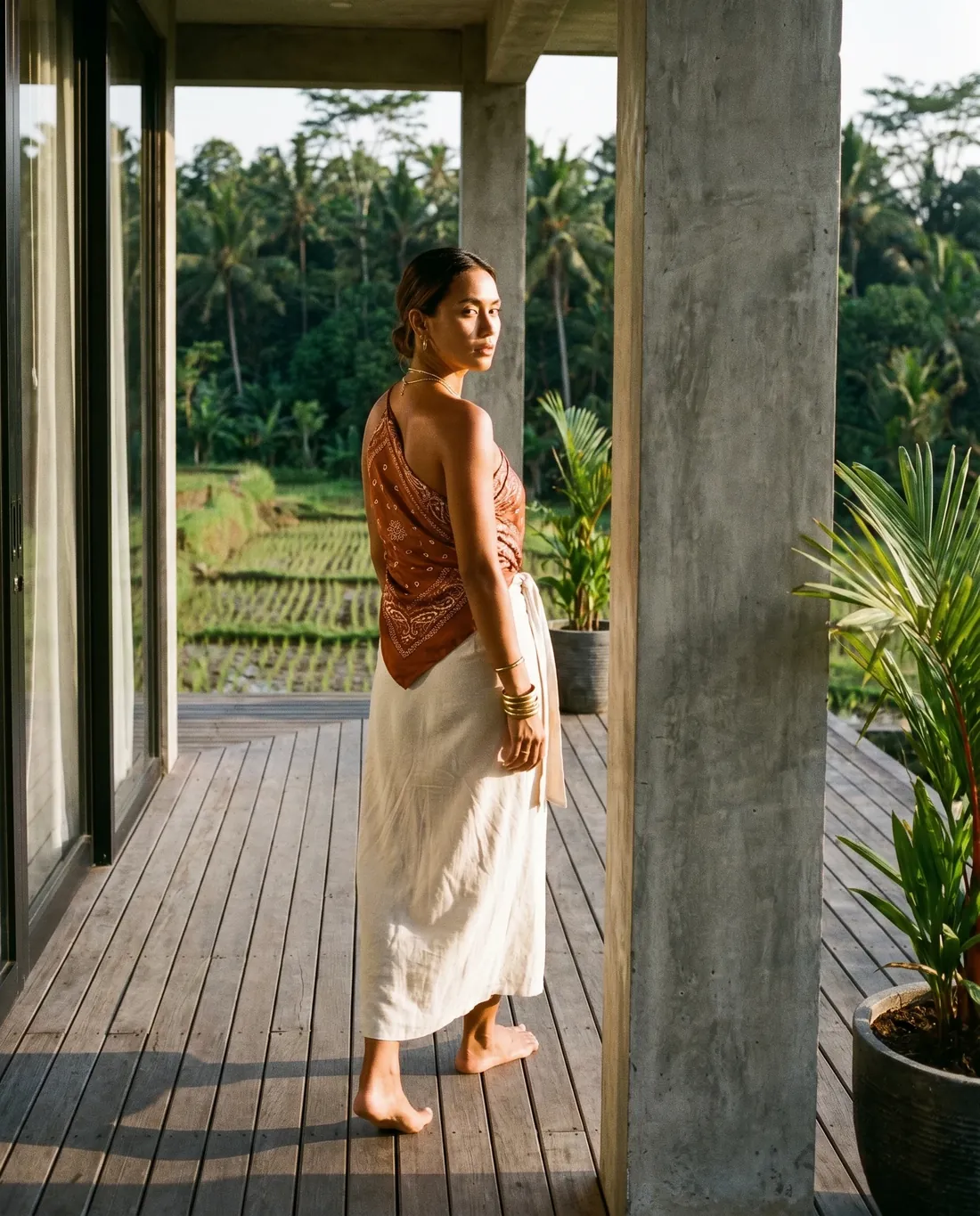 Subject BNE-03 standing on the Villa Soma deck at golden hour. Paisley silk top, cream linen wrap, rice terrace and palms beyond.