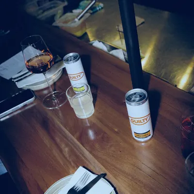 Two GUILTY cans upright on a timber bar at Tintino, beside a glass of ros&eacute; and a place setting.